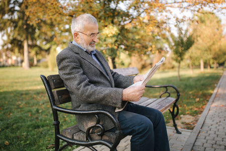 在秋天的公园里,戴着眼镜的大胡子老人在看报纸