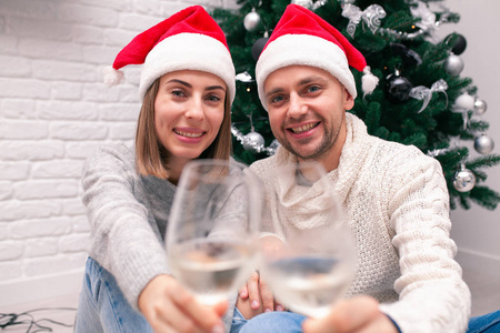 一对幸福的年轻夫妇戴着圣诞帽，在圣诞树旁端着酒杯。新年庆典