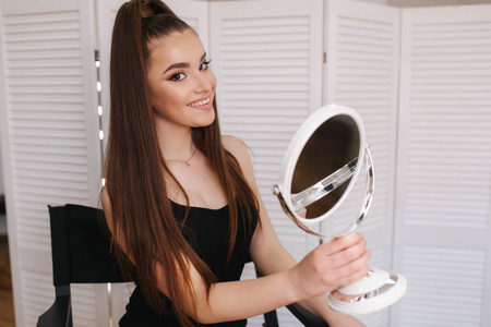 年轻漂亮的模特化妆后照镜子。漂亮的年轻女子坐在美容工作室里。白色折叠式屏风背景