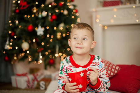 一个穿着圣诞睡衣的小男孩坐在一棵圣诞树旁，手里拿着一杯可可和棉花糖。圣诞节。新年。