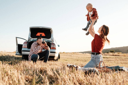 快乐的年轻家庭的妈妈和爸爸带着他们的小儿子享受着夏日周末的野餐，坐在车旁的格子布上，在阳光明媚的日落下，享受假期和公路旅行的概念