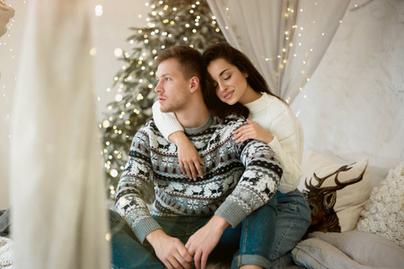 可爱的年轻夫妇漂亮的妻子和英俊的丈夫都穿着温暖的毛衣拥抱在装饰好的沙发上庆祝新年圣诞的喜庆气氛