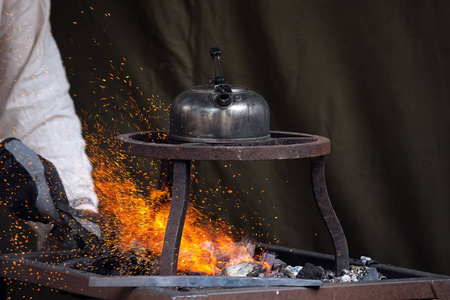铁匠在烧红的煤里加热金属，而茶壶在上面烧开水。巧匠手工锻造金属，红色火花冉冉升起