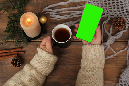 一支蜡烛在木桌上燃烧,智能手机上有空白的绿色屏幕,针织披肩,针叶