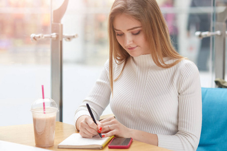 照片中有灵感的迷人的模特在笔记本上写字,独自一人在咖啡馆里,桌上放