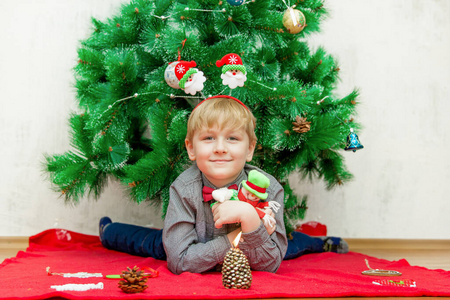 快乐的微笑着的浅色头发的小男孩躺在圣诞树下的红色毯子上，周围有圣诞装饰品