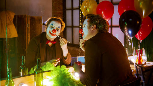 阴险的笑容小丑在化妆间里,一个男人在镜子前把脸涂上小丑画照片