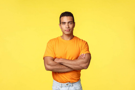 自信上进的帅哥，健美运动员穿着休闲橙色t恤，双臂交叉放在胸前，看上去自信而坚定，微笑愉快，邀请加入健身俱乐部，站在黄色背景