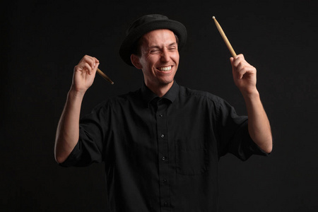 穿着黑色衬衫和帽子的时髦男子鼓手，在深色背景上用棍子打鼓。
