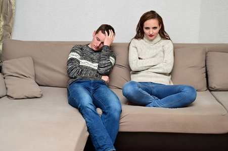 家庭冲突的概念。一个年轻的不幸家庭的肖像愤怒的黑发女孩和不高兴的家伙在家里的沙发上穿着毛衣。不满的妻子责骂丈夫。