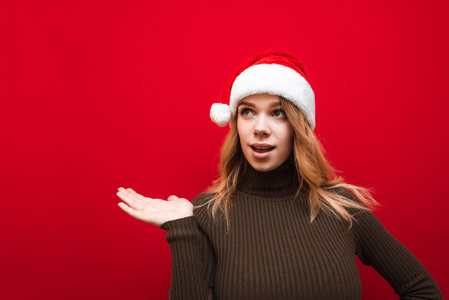 圣诞帽上穿着温暖毛衣的迷人女孩惊讶地看着圣诞老人，用手指着那片与世隔绝的空旷空间。情绪激动的女孩手里拿着红色背景的版面。