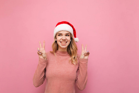 快乐女孩穿着粉色毛衣，头戴圣诞老人帽，戴着无线耳机，站在粉红色的背景上，看着空空荡荡的座位，用手指和微笑表示和平的姿态。孤立的。