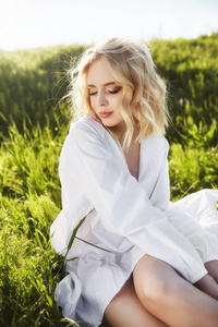 Girl in a long white dress sits on the grass in a field. Blonde 