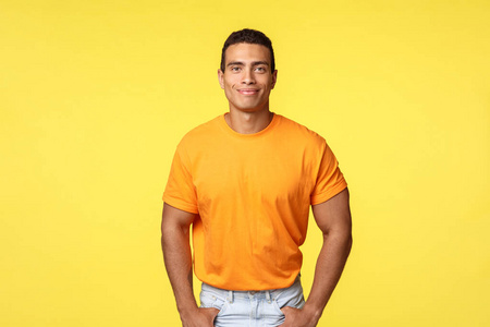 阳刚时尚的现代男士，穿着橙色t恤，手插口袋，微笑友好，来帮助邻居，随意闲聊，站在快乐的黄色背景下