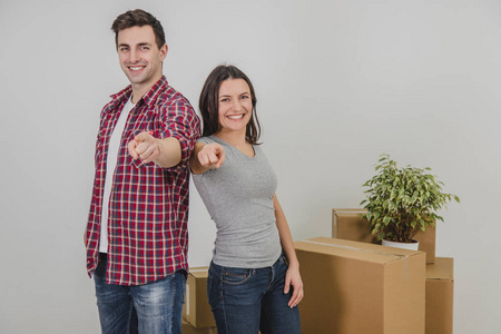 幸福的男人和女人肩并肩站在他们新公寓的纸箱旁边，用手指指着相机，微笑着。
