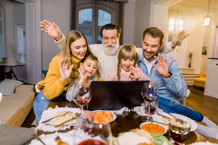 美丽的一家人，坐在餐桌边吃晚饭，微笑着，开心地用着笔记本电脑。家人在家用笔记本电脑打视频电话庆祝