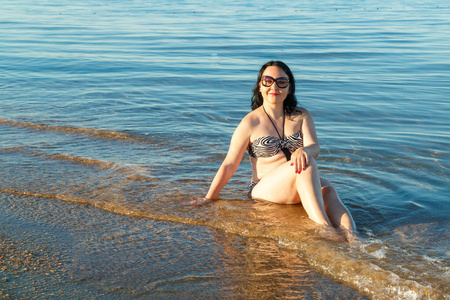 一个黑发女人穿着泳衣戴着眼镜坐在海边。