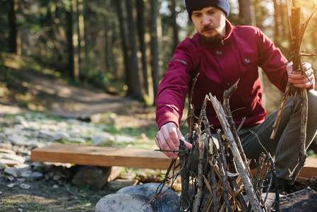 一个穿红夹克的年轻人在森林里点燃篝火。旅人坐在篝火旁。探索真正的野外生活方式
