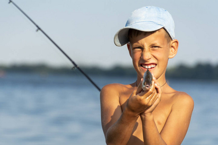 快乐的微笑男孩手里拿着拿着的淡水鱼在鱼竿和河流的背景下