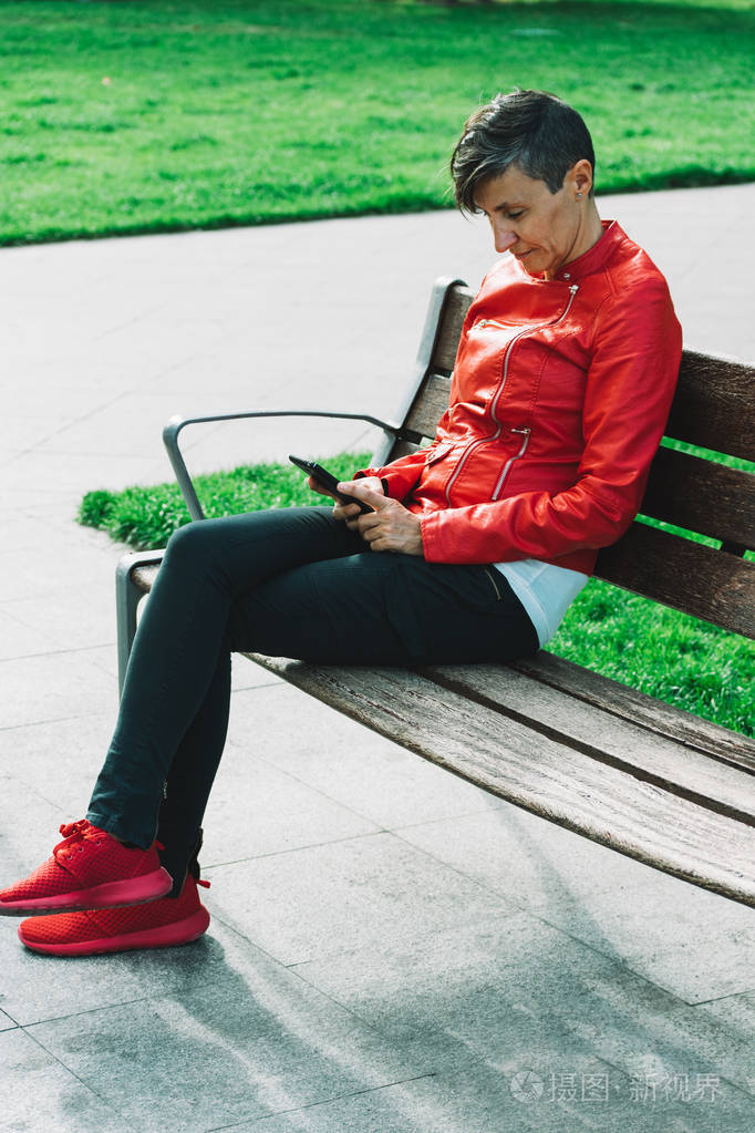 穿着红夹克的女人坐在长椅上拿着智能手机