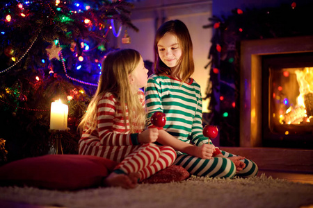 圣诞夜，在一个舒适黑暗的客厅里，快乐的小姐妹们一起在壁炉旁吃着覆盖着糖霜的红苹果。在家庆祝圣诞节。