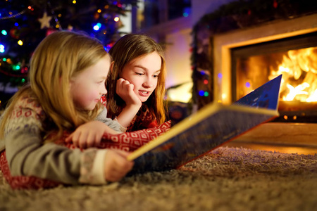 平安夜，在一个舒适黑暗的客厅里，快乐的小姐妹们在壁炉旁一起读故事书。在家庆祝圣诞节。