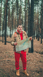 一个在树林里拿着地图的旅游者。一个英俊的男人背着一个背包，在寒冷的天气里站在一棵树旁。
