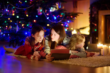 圣诞夜，母亲和两个可爱的小女儿在温暖舒适的客厅壁炉旁用平板电脑。