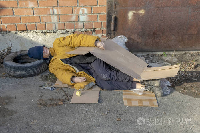 一个无家可归的女人穿着一件破旧的黄色夹克，戴着一顶蓝色的帽子，躺在人行道上的纸板上睡觉
