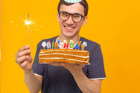 一个有趣的正面人物的画像，戴着一顶纸帽子，戴着眼镜，手里拿着一个自制的祝贺蛋糕，背景是黄色。概念，乐趣和庆祝。