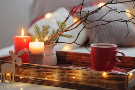 Red cup of tea on tray with burning candles on background sofa 