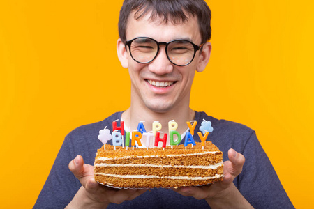 一个疯狂开朗的亚洲年轻人，戴着眼镜，手里拿着一支点燃的蜡烛，黄色背景的自制蛋糕表示祝贺。生日和周年庆祝概念。