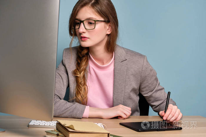 一个穿着灰色夹克戴着眼镜的辫子的女孩坐在桌子旁，看着显示器，在制作图形平板电脑。