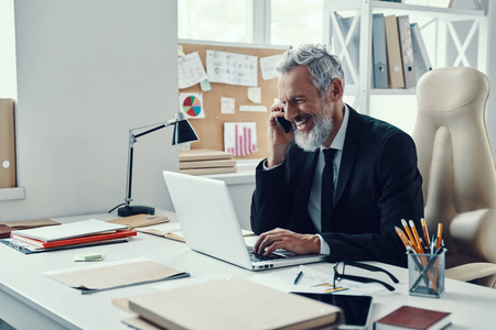 在现代办公室工作时，穿着正装的快乐成熟男士使用笔记本电脑和打电话