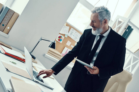 在现代办公室工作时，体贴成熟的男士正装，使用笔记本电脑和数字平板电脑