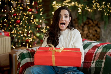 年轻漂亮的女士穿着温暖的毛衣拿着礼物坐在沙发上庆祝新年和圣诞节，看起来惊讶的节日气氛