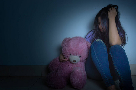 一个悲伤的女人抱着她的膝盖哭着坐在黑暗中的泰迪熊