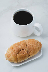 White mug with black coffee and croissant on white stone table. 