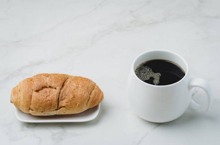 White mug with black coffee and croissant on white stone table. 