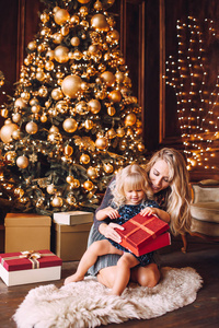 冬天，在舒适的起居室里，年轻的金发碧眼的母亲和穿着闪亮衣服的小女儿在圣诞树旁打开圣诞礼物