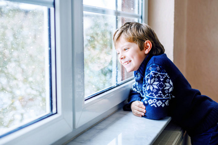 圣诞节或早上，快乐可爱的小男孩坐在窗边看着外面的雪。笑容可掬的健康孩子着迷于观察降雪和大雪花