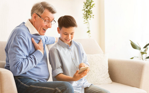 坐在室内沙发上教爷爷使用智能手机的男孩
