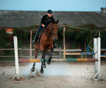 Horsewoman riding on brown horse and jumping the fence in sandy 