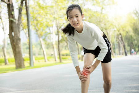 不开心的亚洲小女孩在阳光明媚的日子玩耍时，腿部疼痛或小腿肌肉受伤，女性青少年双手握住双腿，夏季运动时感到膝盖疼痛，身体受伤概念