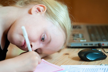 一个低头写字的金发女孩.背景中的笔记本电脑和鼠标不对焦照片
