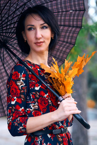 美丽优雅的女子站在秋天的城市公园里，拿着一束黄叶和一把伞