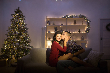 幸福，在黑暗中相爱的情侣，在沙发上的夜晚，黑暗的背景，圣诞树的灯光。圣诞夜。新年。拥抱和亲吻。吸引人，浪漫的夜晚和约会。