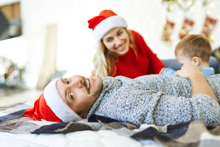 照片中，英俊的微笑男子戴着红色圣诞帽，躺在床上，儿子和妻子。