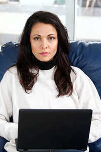 穿着毛衣的黑发女人坐在沙发上，手里拿着笔记本电脑