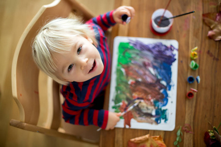 Sweet toddler boy with red apron, painting at home 